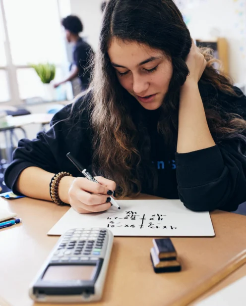 A student with long brown hair writes math equations on a whiteboard at a desk, with a calculator and eraser nearby in a classroom setting.