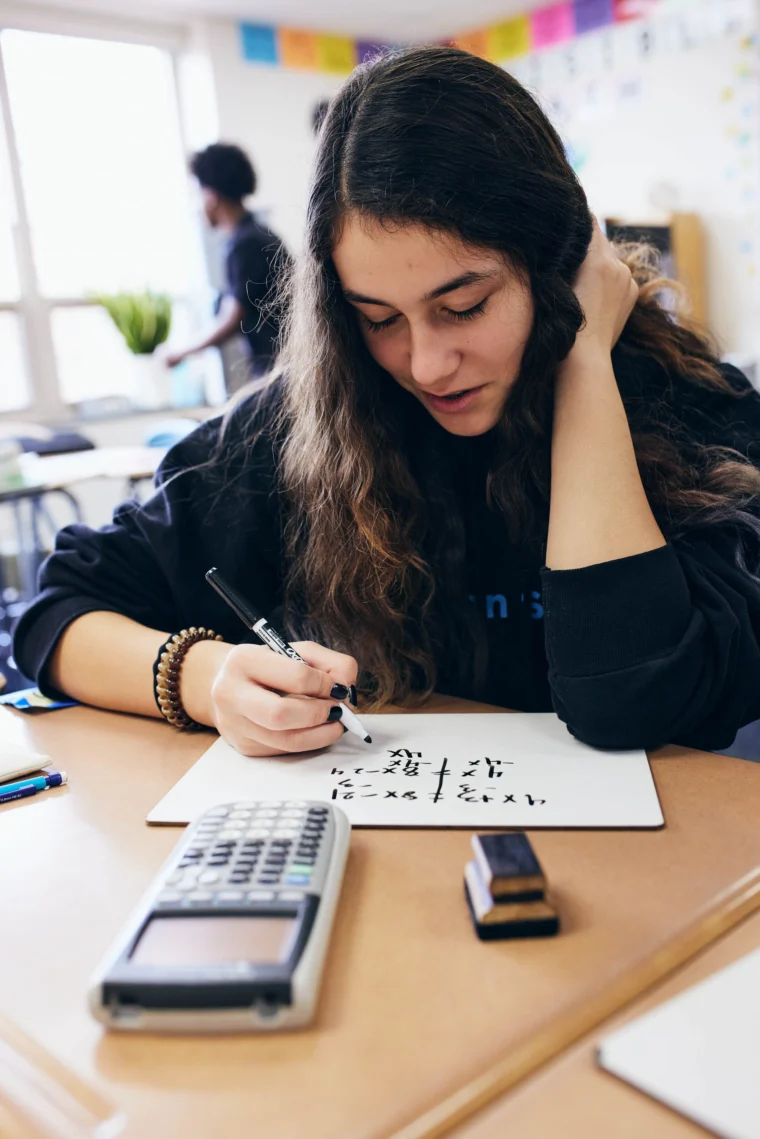 A student with long brown hair writes math equations on a whiteboard at a desk, with a calculator and eraser nearby in a classroom setting.