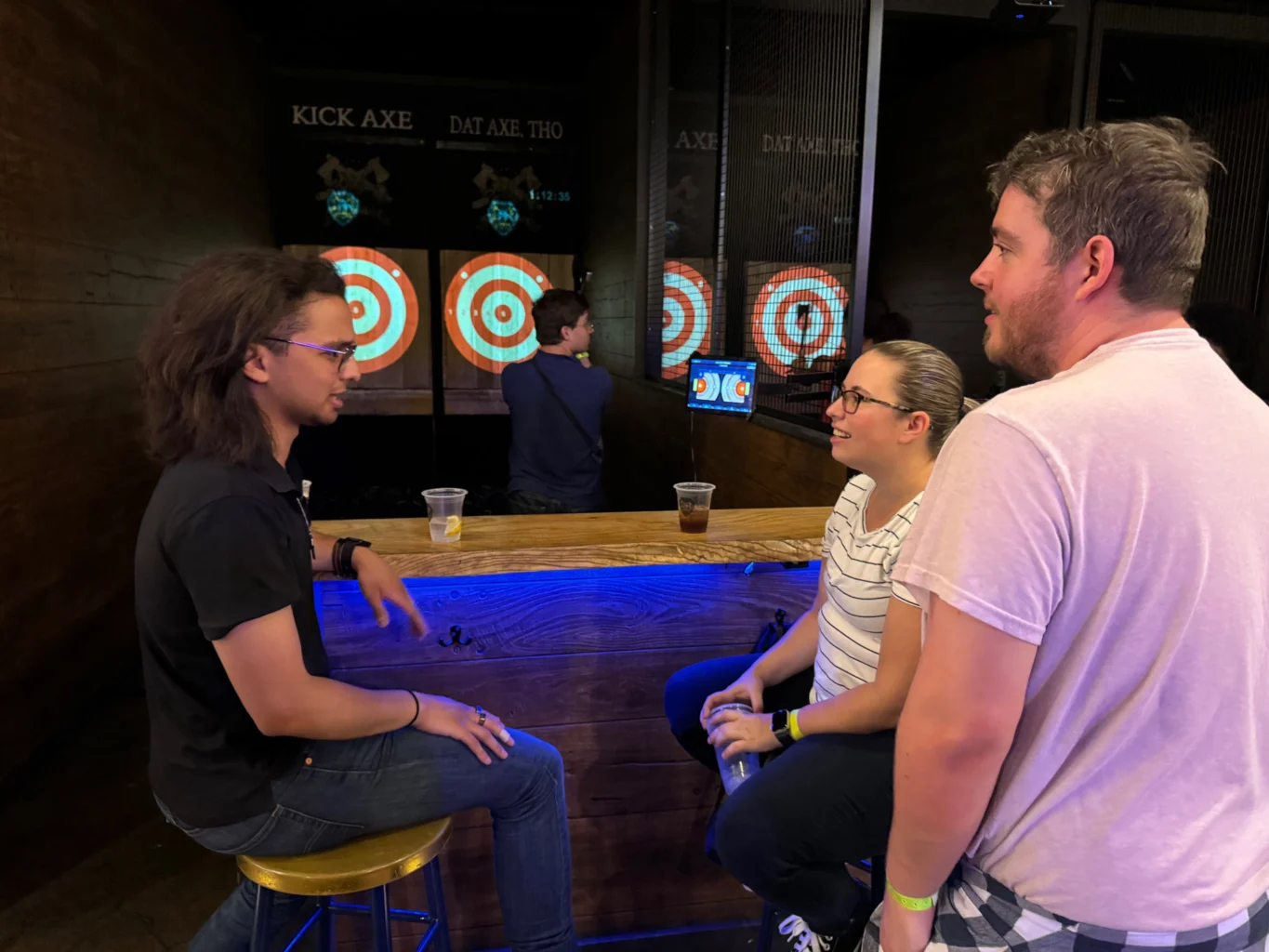 Three people converse at a bar table while another person throws axes at target boards in the background at an indoor axe-throwing venue.