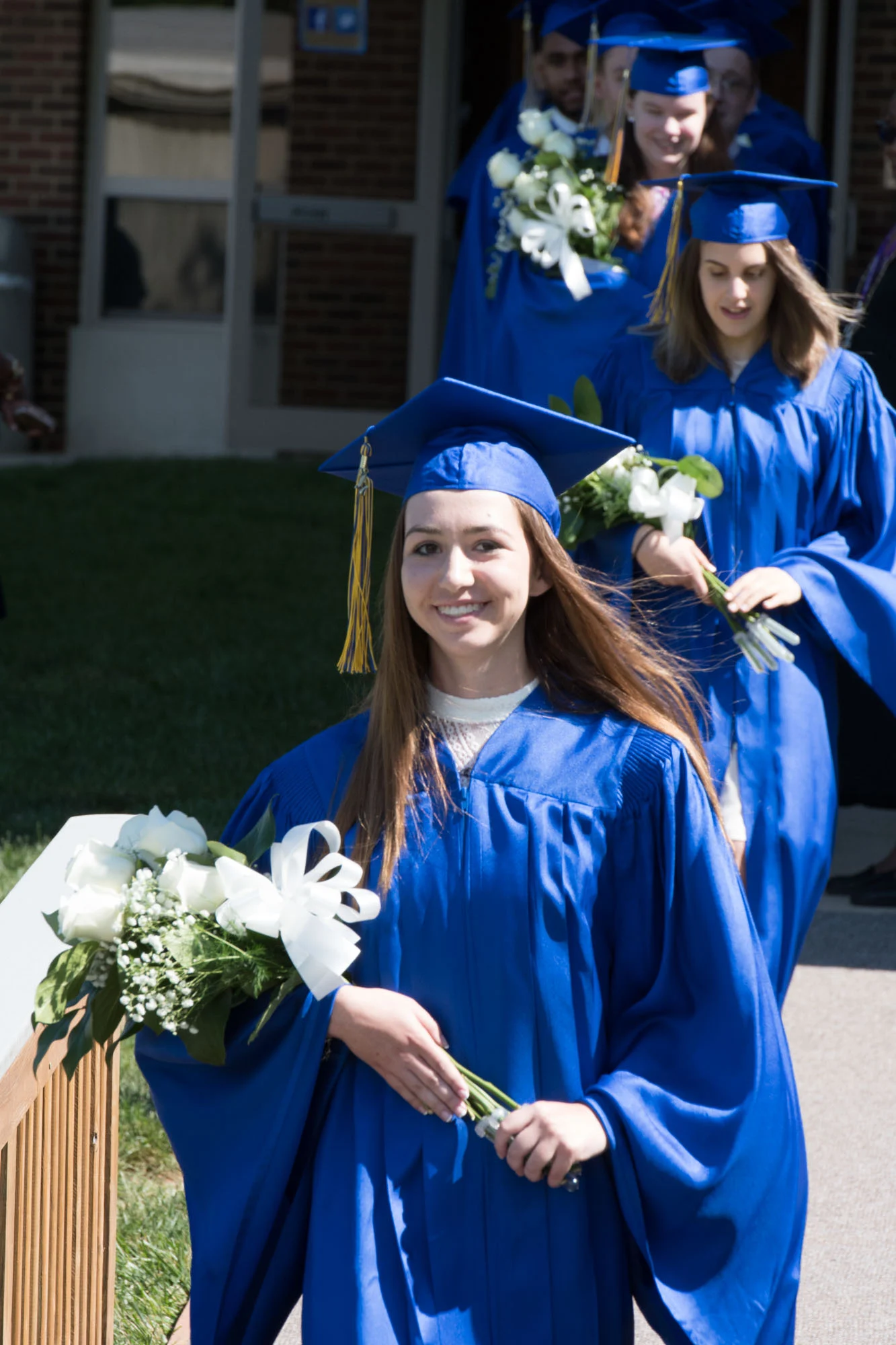 Several graduates in blue caps and gowns walk outside in a line, holding bouquets of white flowers and greenery, during a graduation ceremony.