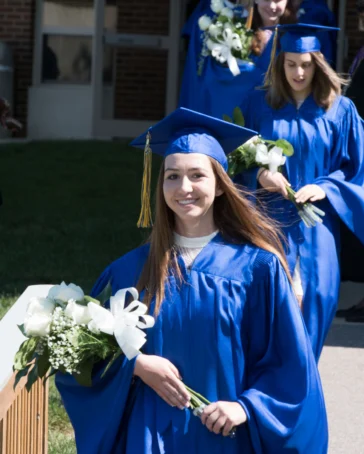 Several graduates in blue caps and gowns walk outside in a line, holding bouquets of white flowers and greenery, during a graduation ceremony.