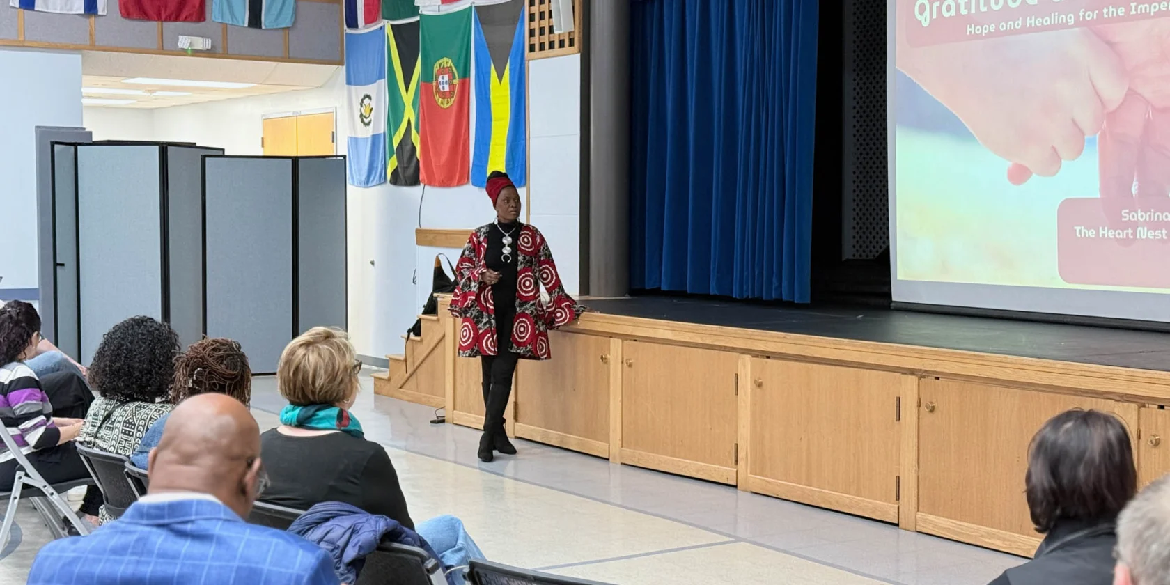 A speaker stands on stage in front of an audience, with international flags overhead and a presentation titled “Gratitude and Forgiveness” displayed on the screen.