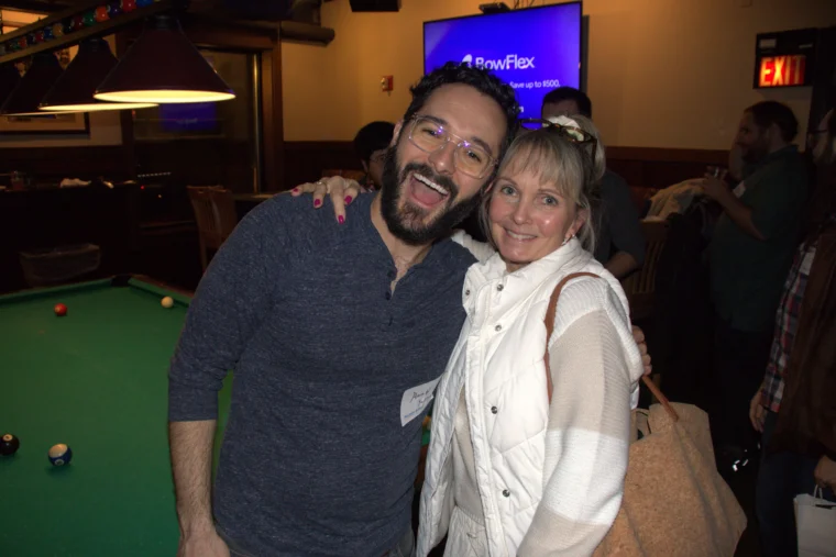 A man and woman smile at the camera in front of a pool table at an indoor social gathering. The man has his arm around the woman. People are visible in the background.