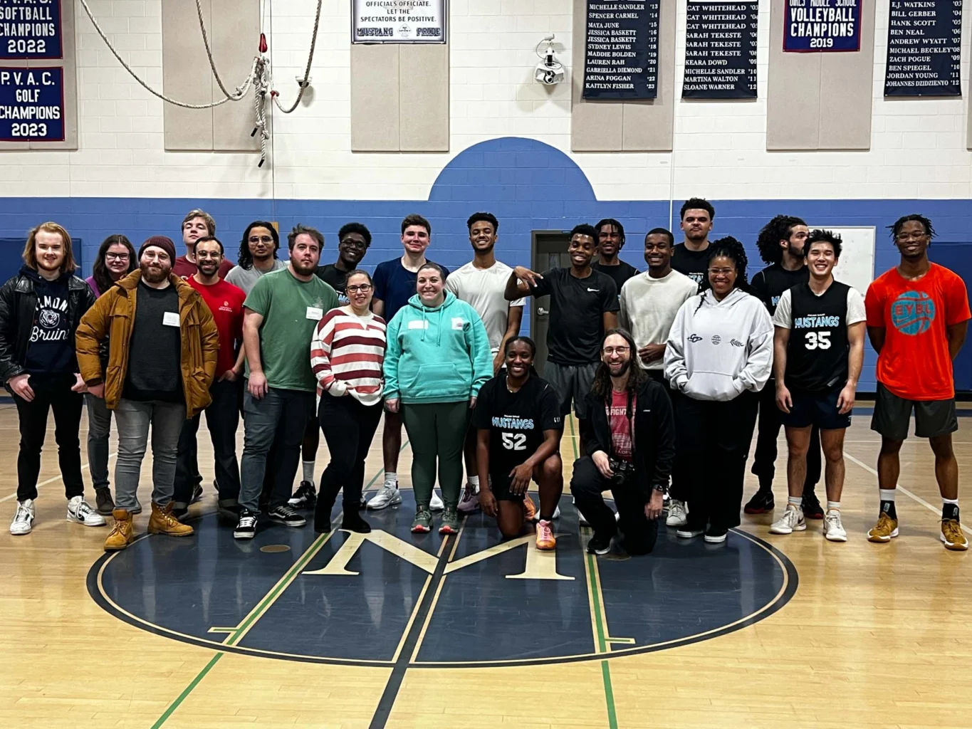 A group of 23 people pose for a photo on a gymnasium floor with banners and a basketball hoop visible in the background.