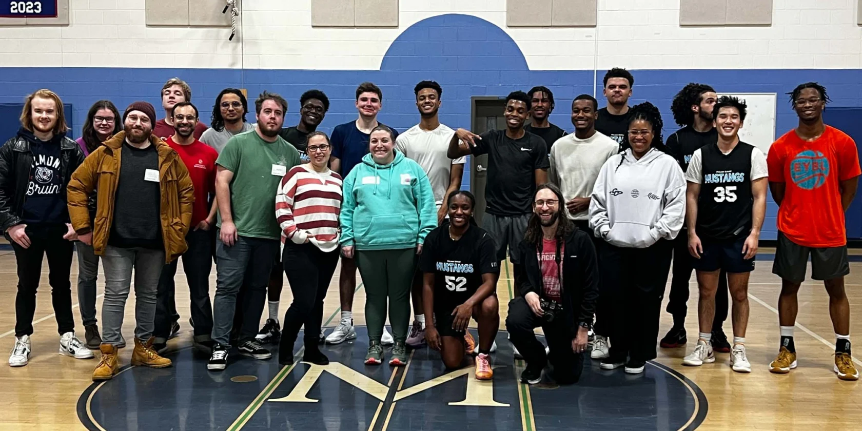 A group of 23 people pose for a photo on a gymnasium floor with banners and a basketball hoop visible in the background.