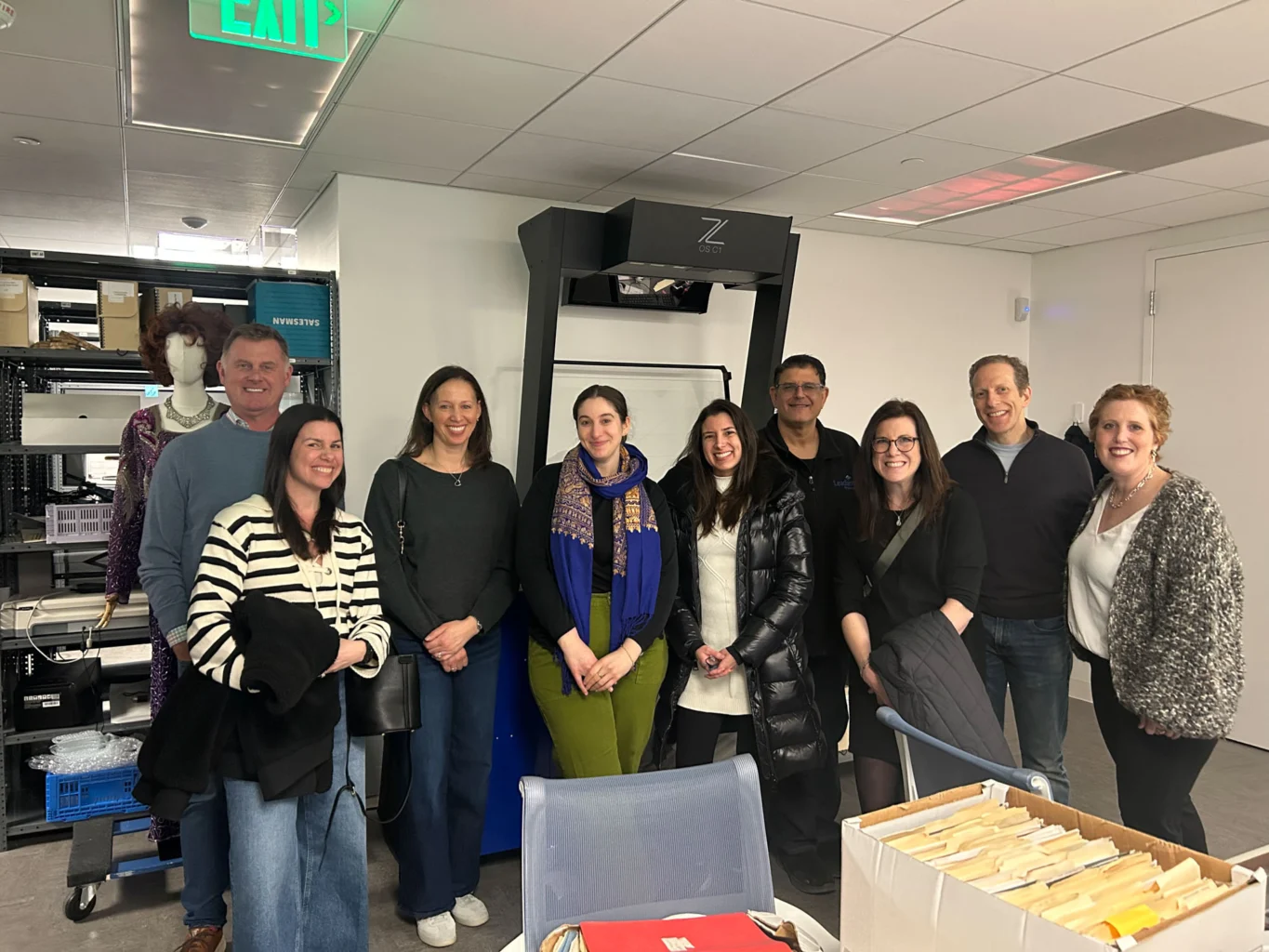 A group of nine people pose for a photo in an office space with shelves, equipment, and file folders visible in the background.