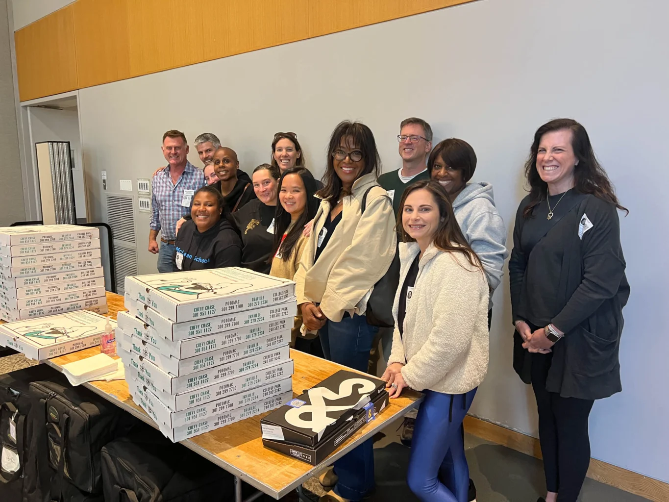 A group of people stands and smiles behind a table stacked with pizza boxes and swag items in a well-lit room.