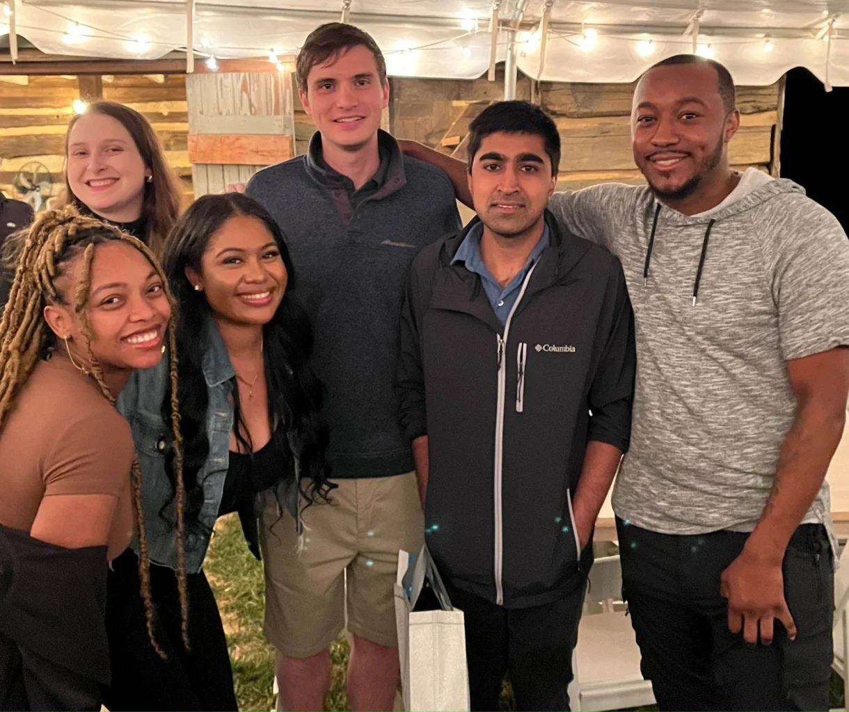 Six adults pose together and smile for a group photo at an outdoor event under string lights.