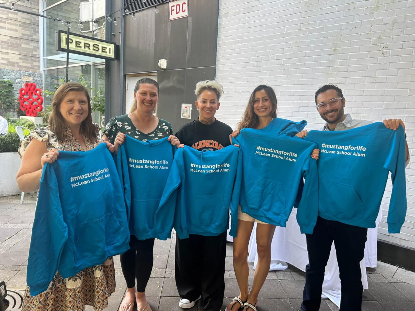 Five people stand outside holding blue "#mustangforlife McLean School Alum" sweatshirts and smiling at the camera.