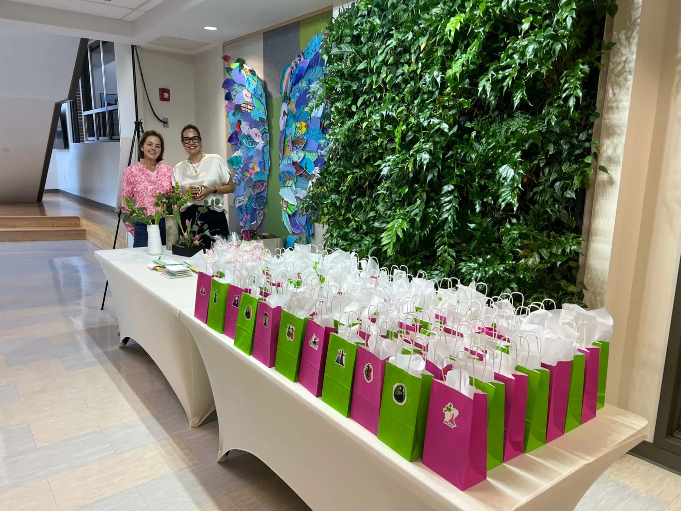 Two people stand behind a table covered with green and pink gift bags, in a hallway decorated with a vertical garden and colorful artwork.