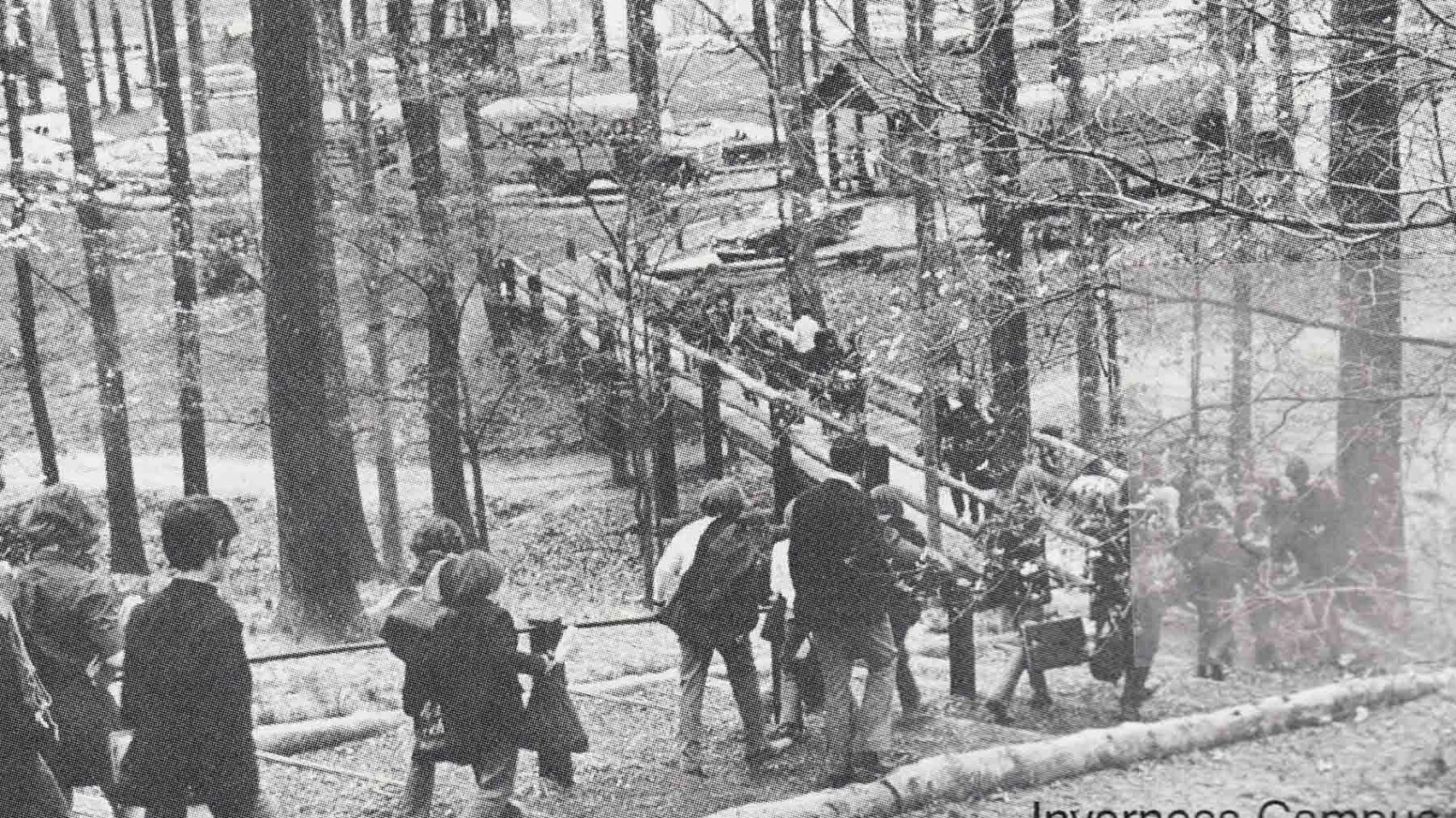 Black and white photo of people walking along a wooded path and bridge on the Inverness Campus, with parked cars visible in the background.