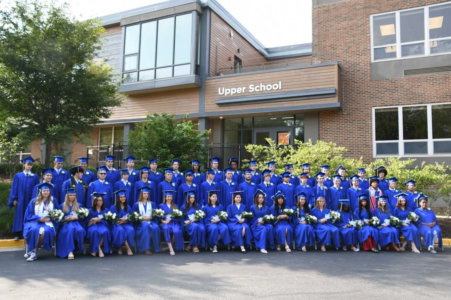 A large group of students in blue graduation gowns and caps pose for a photo in front of a building labeled "Upper School.