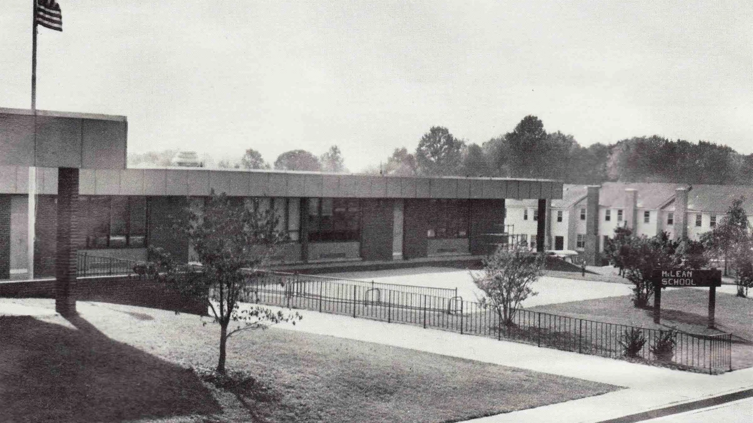 Black and white photo of a low, single-story school building with an American flag and a sign reading "McLean School" near a row of houses and trees.