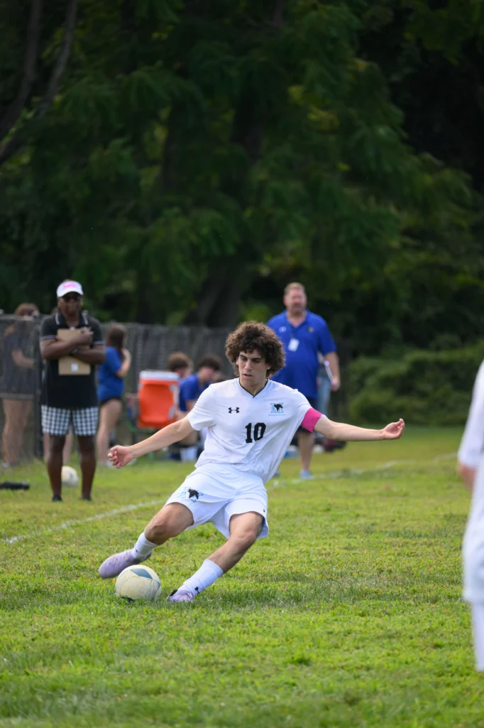 A soccer player in a white uniform with the number 10 is sliding to kick the ball on a grassy field, with teammates and spectators in the background.