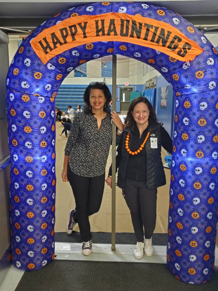 Two women stand under a blue archway decorated with skulls and pumpkins, with a sign that reads "Happy Hauntings" in a gymnasium setting.