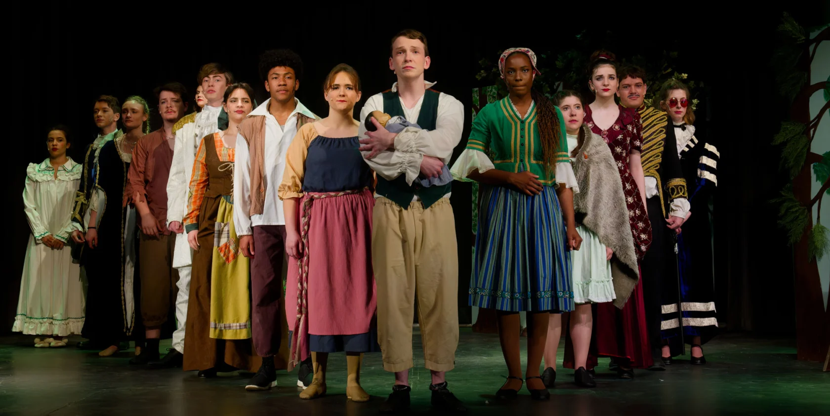 A group of actors in period costumes stand in formation on stage, facing forward, under theatrical lighting.