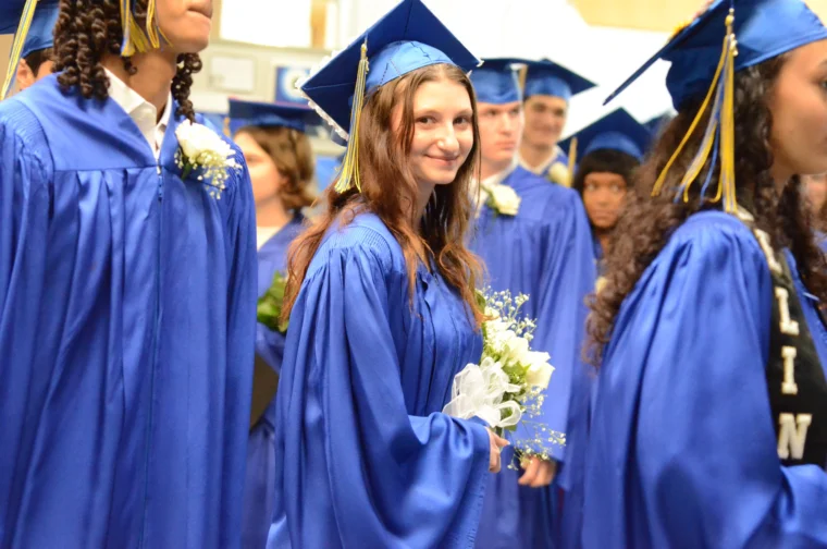 Students wearing blue graduation gowns and caps stand in a group; a young woman in the center holds a bouquet and smiles at the camera.