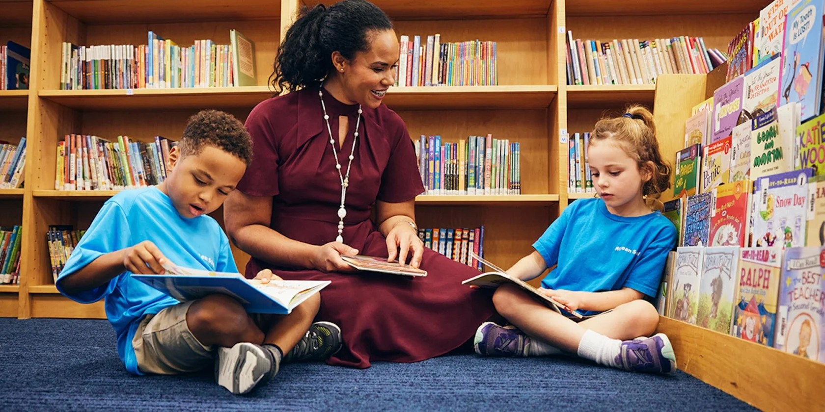 A woman sits on the floor in a library with two children, all reading books together near shelves filled with various children's books.