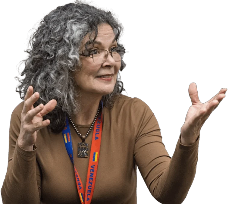 A woman with curly gray hair wearing glasses, a brown top, and a "Venezuela" lanyard gestures with both hands while speaking.