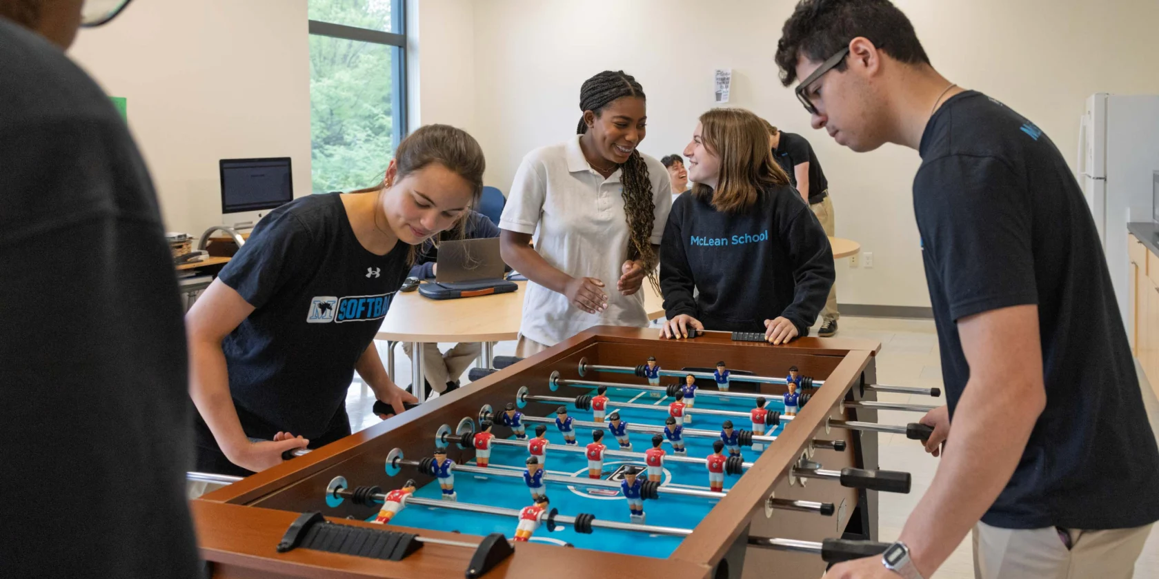 Five young people play foosball in a bright room with large windows, computers, and a fridge in the background.