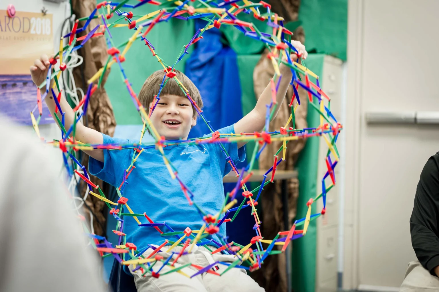 A boy in a blue shirt sits and holds a large expandable geometric toy, smiling, in a brightly lit indoor setting.