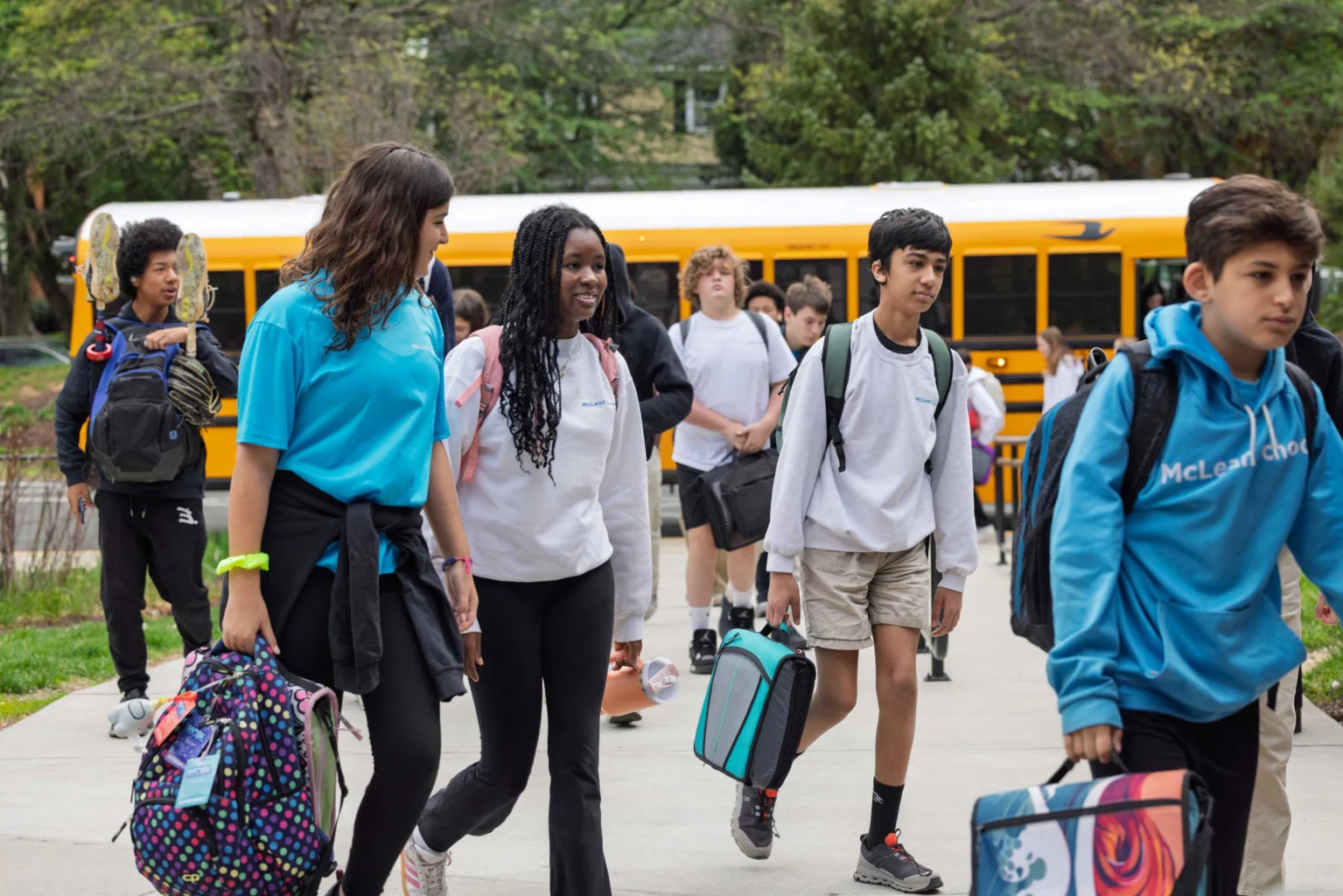 Students walk on a sidewalk carrying backpacks and lunch bags, with a yellow school bus and trees in the background.