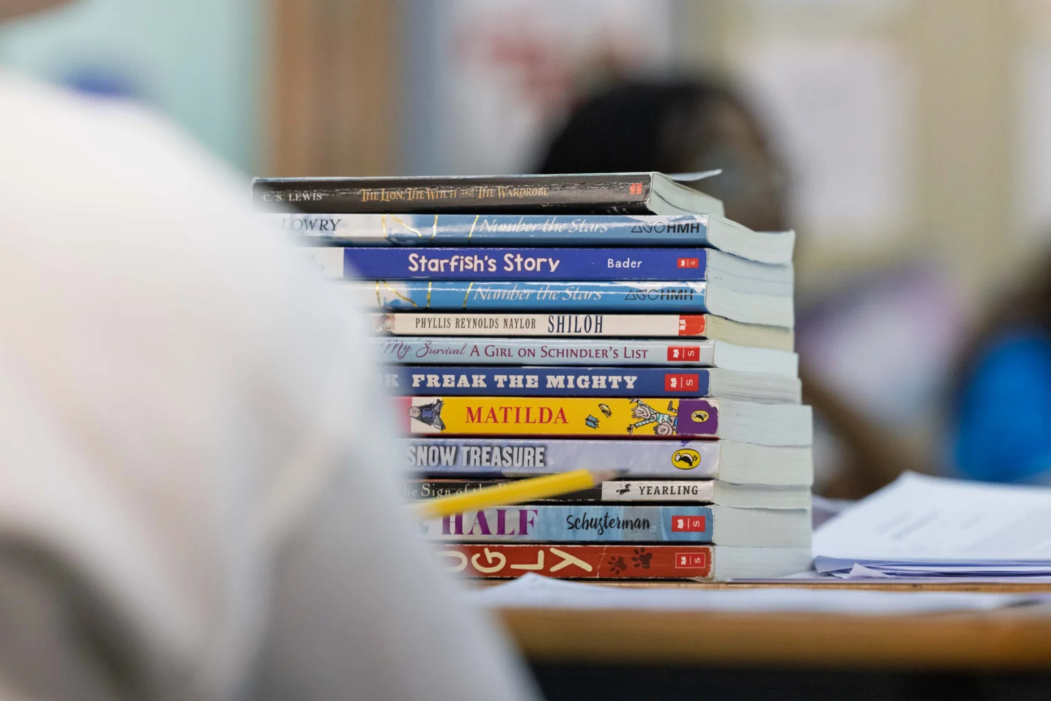 A stack of assorted children's and young adult books is placed on a table, with blurred figures and a classroom setting in the background.