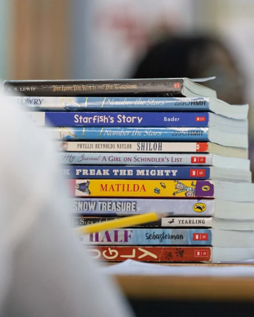 A stack of assorted children's and young adult books is placed on a table, with blurred figures and a classroom setting in the background.