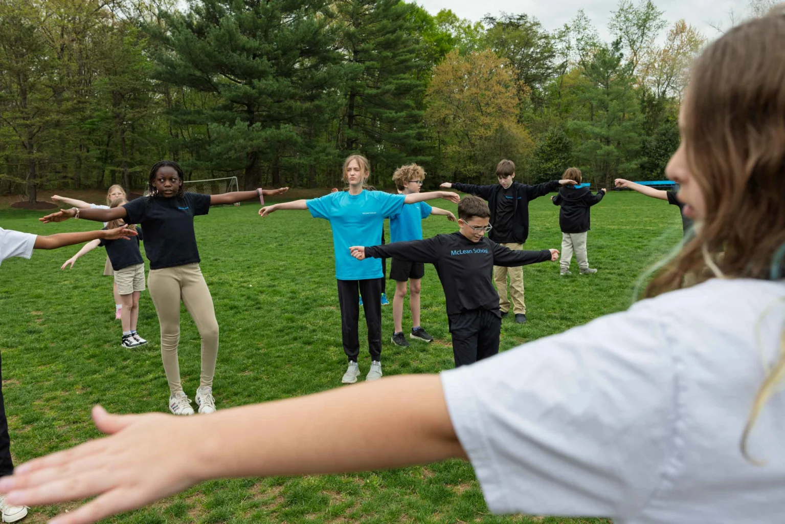 A group of students stands in a circle outside on grass with arms outstretched, participating in an outdoor activity.