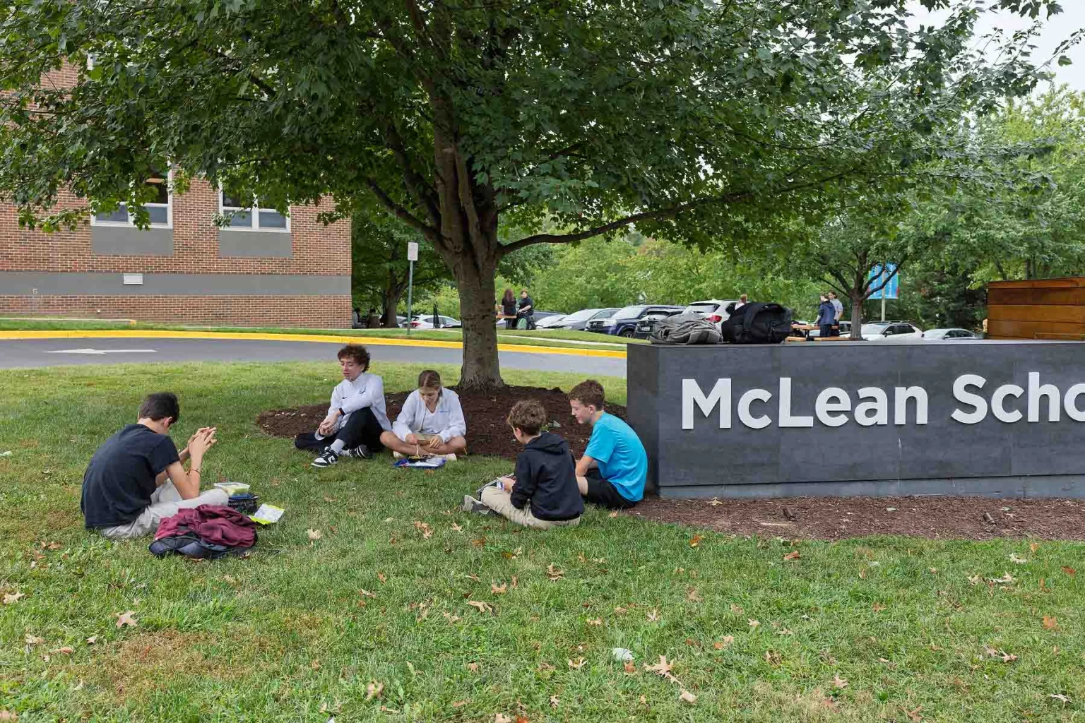 Five students sit on the grass near a tree by a large McLean School sign, talking and writing, with backpacks nearby and a building in the background.