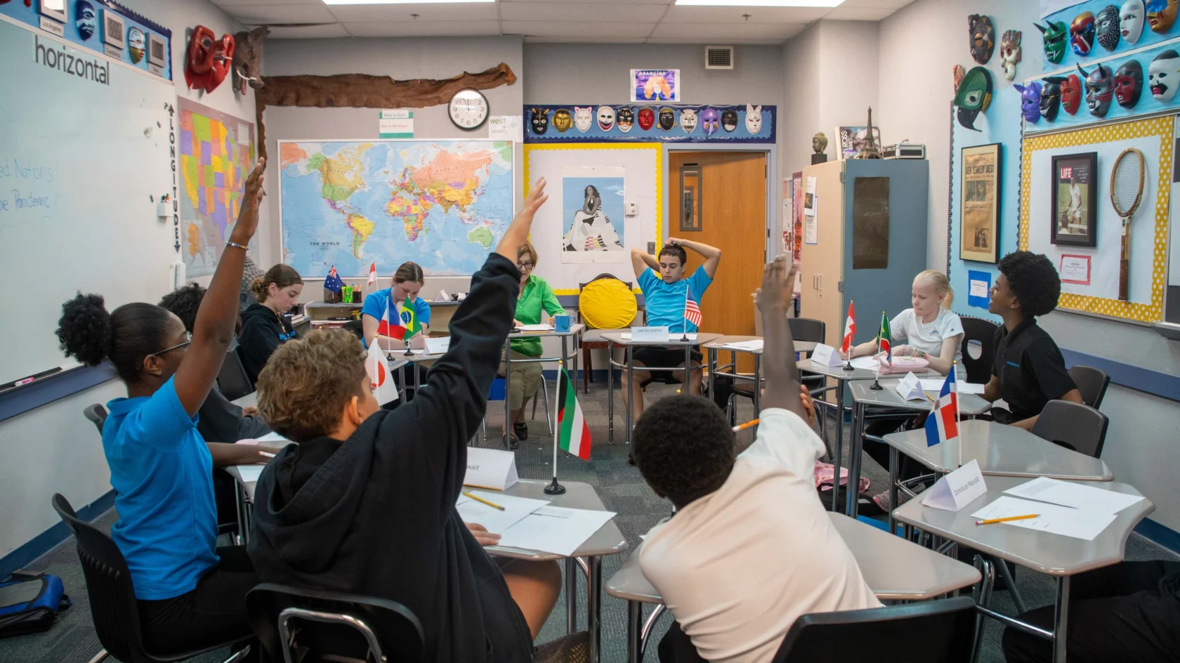 Students sitting in a classroom arranged in a circle raise their hands, with international flags on desks and educational posters on the walls.