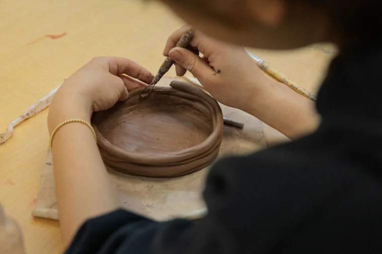 A person shapes a round clay bowl with a sculpting tool on a wooden table.