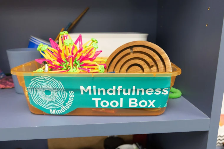 A plastic bin labeled "Mindfulness Tool Box" contains sensory toys and wooden objects, placed on a gray shelf.