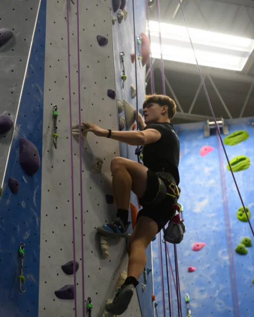 Person wearing a harness climbs an indoor rock wall, gripping holds and using ropes for safety under bright overhead lights.
