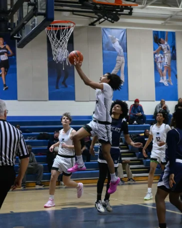 A basketball player in a white uniform jumps toward the hoop for a layup while players in dark uniforms attempt to defend in a gymnasium.