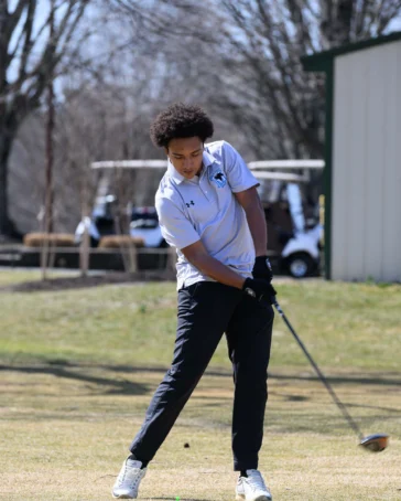 A person wearing a white polo and black pants swings a golf club on a golf course, with trees and golf carts in the background.
