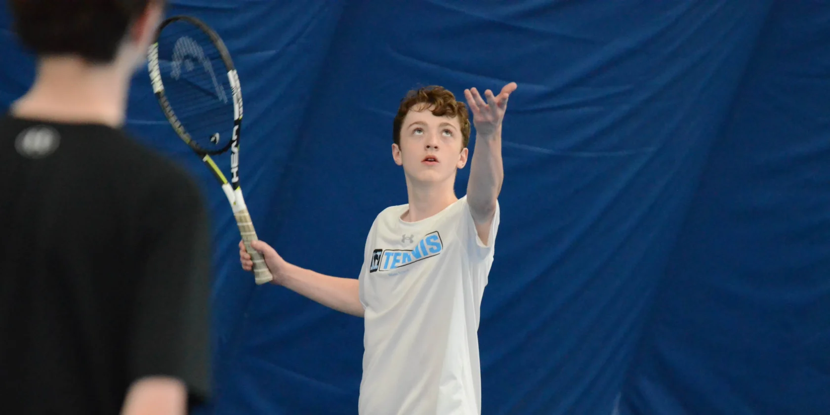 A boy in a white shirt prepares to serve a tennis ball indoors, with another person visible in the foreground.