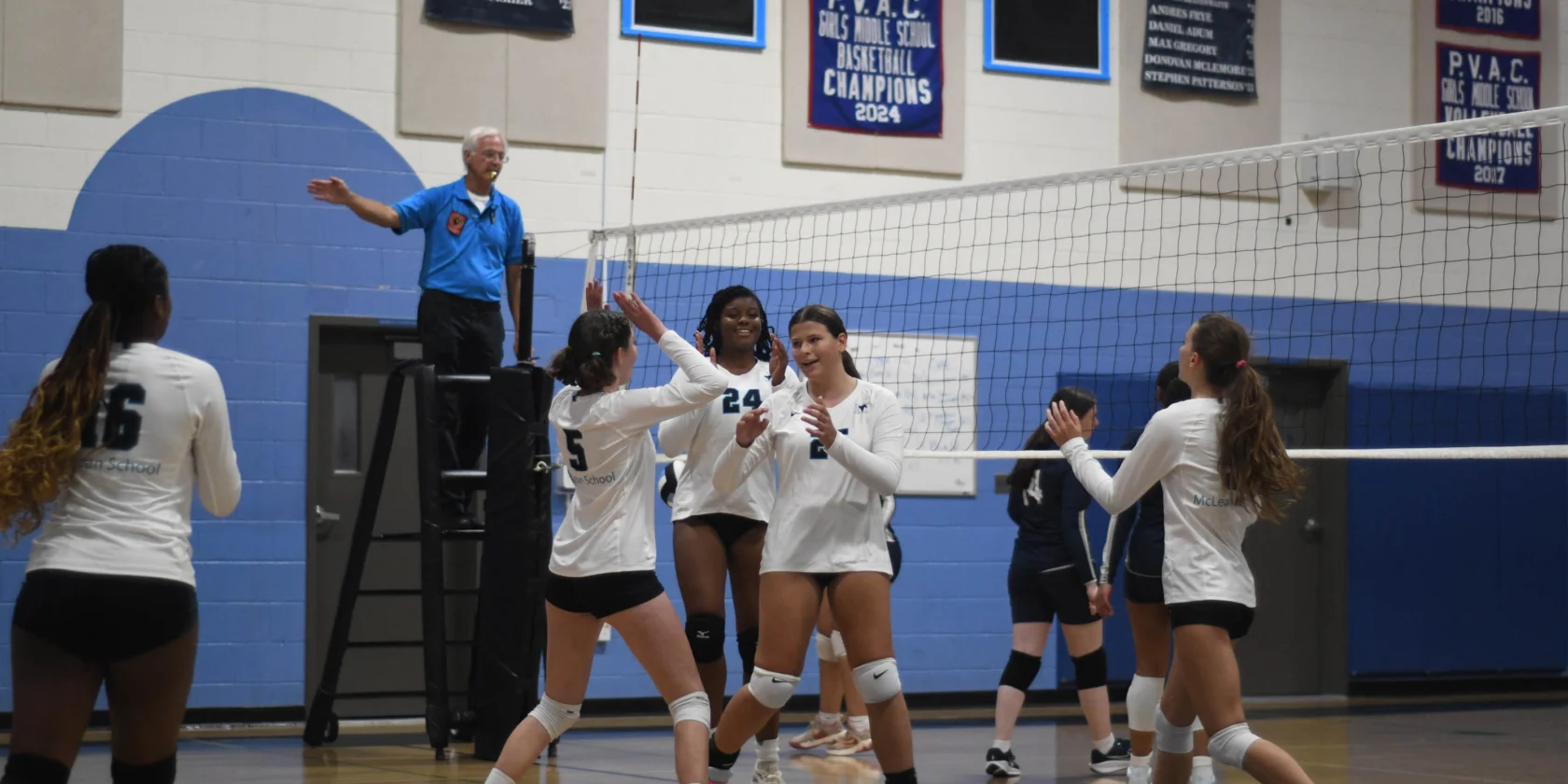 A girls’ volleyball team in white jerseys celebrates a point on an indoor court as a referee stands on the officiating stand.