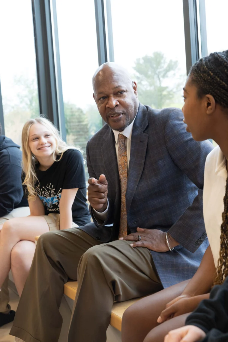 A man in a suit sits on a bench, talking and gesturing to two young women, one smiling, in front of large windows.