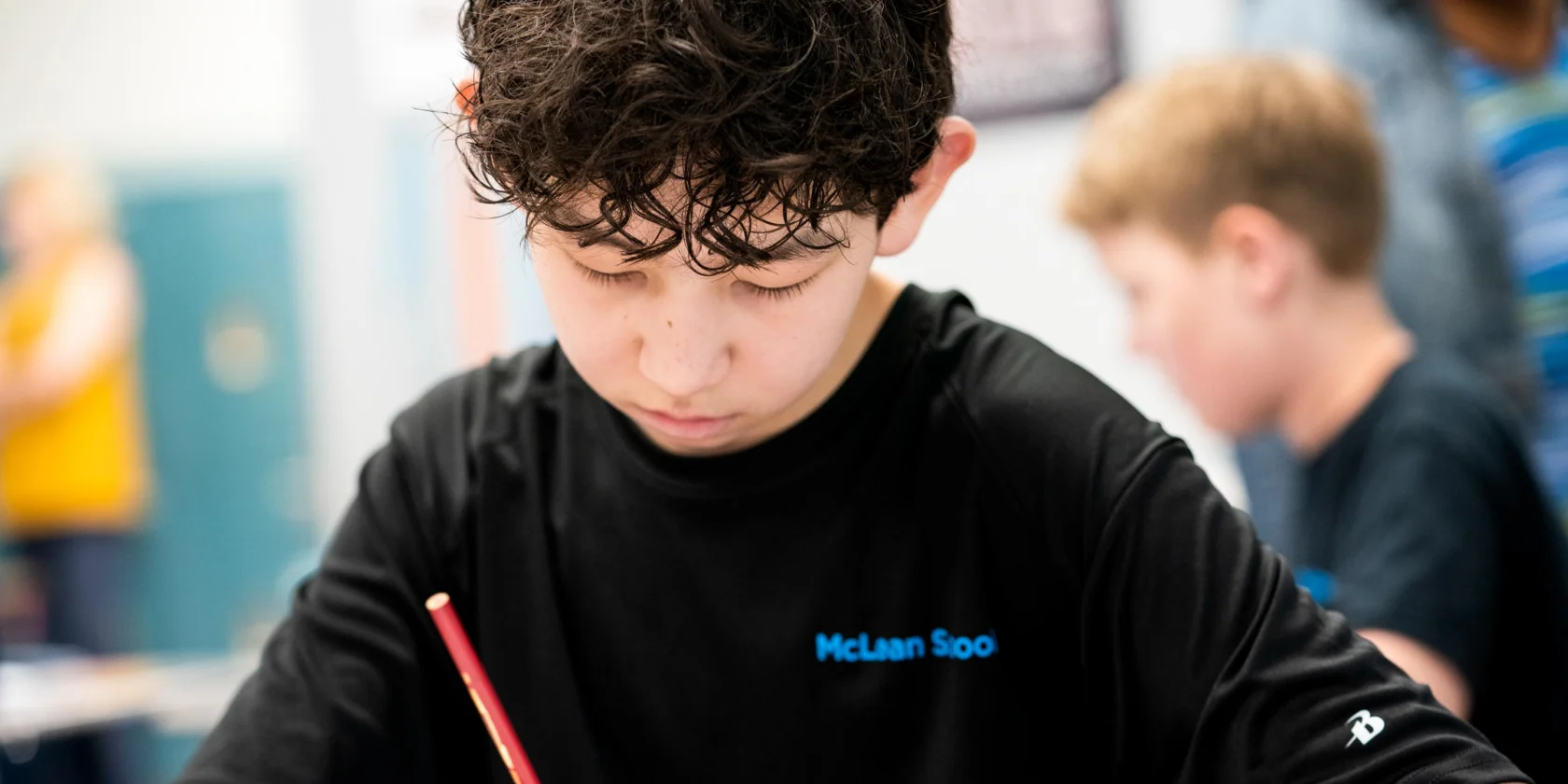 A boy with curly hair writes in a notebook with a red pencil at a desk, while another student works in the background.