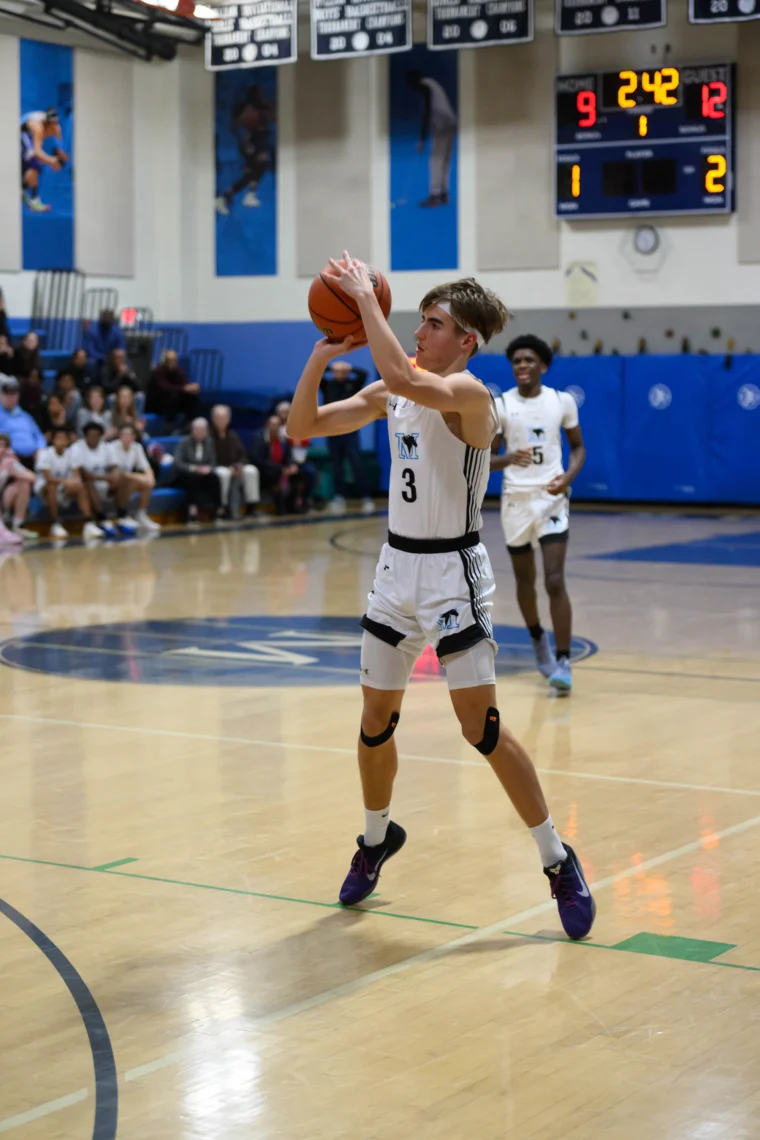 A basketball player in a white uniform prepares to shoot the ball during a game in a gym, with spectators and a scoreboard in the background.