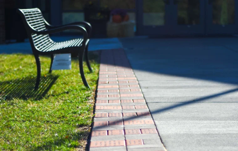A metal bench sits on grass beside a walkway lined with engraved bricks, with building doors visible in the background.