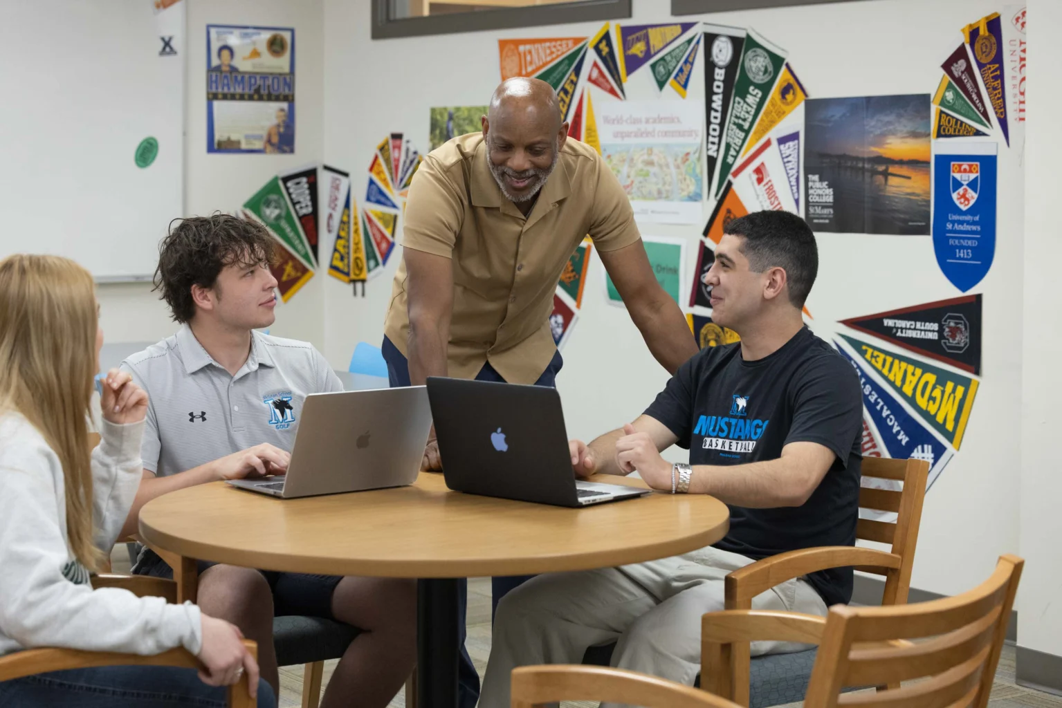 A group of students sit around a table with laptops while an instructor stands and talks to them in a classroom decorated with college pennants.