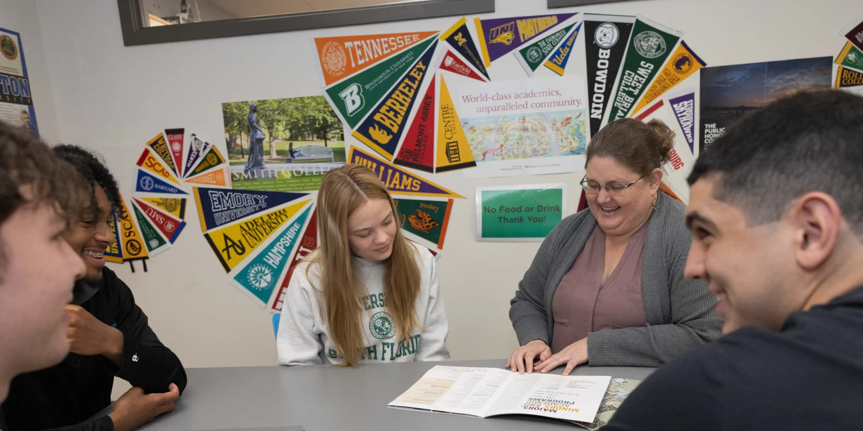 Five people sit around a table in an office decorated with college pennants, looking at papers and a laptop, with one person leading the discussion.