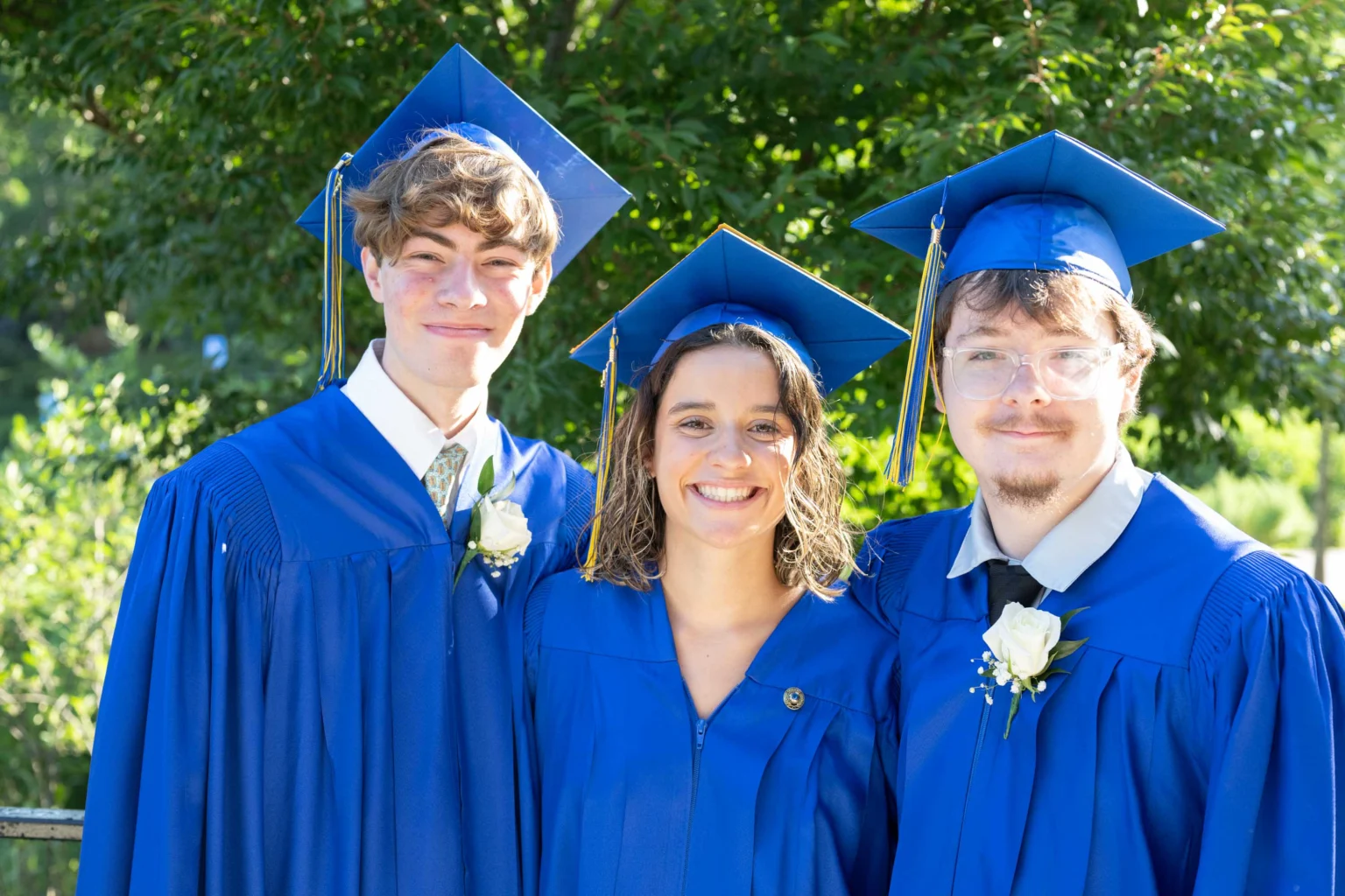 Three graduates wearing blue caps and gowns stand outdoors, smiling at the camera with greenery in the background.