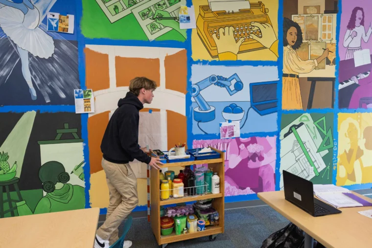 A student pushes a cart with art supplies in front of a colorful classroom mural featuring illustrations of artists, musicians, writers, and scientific equipment.