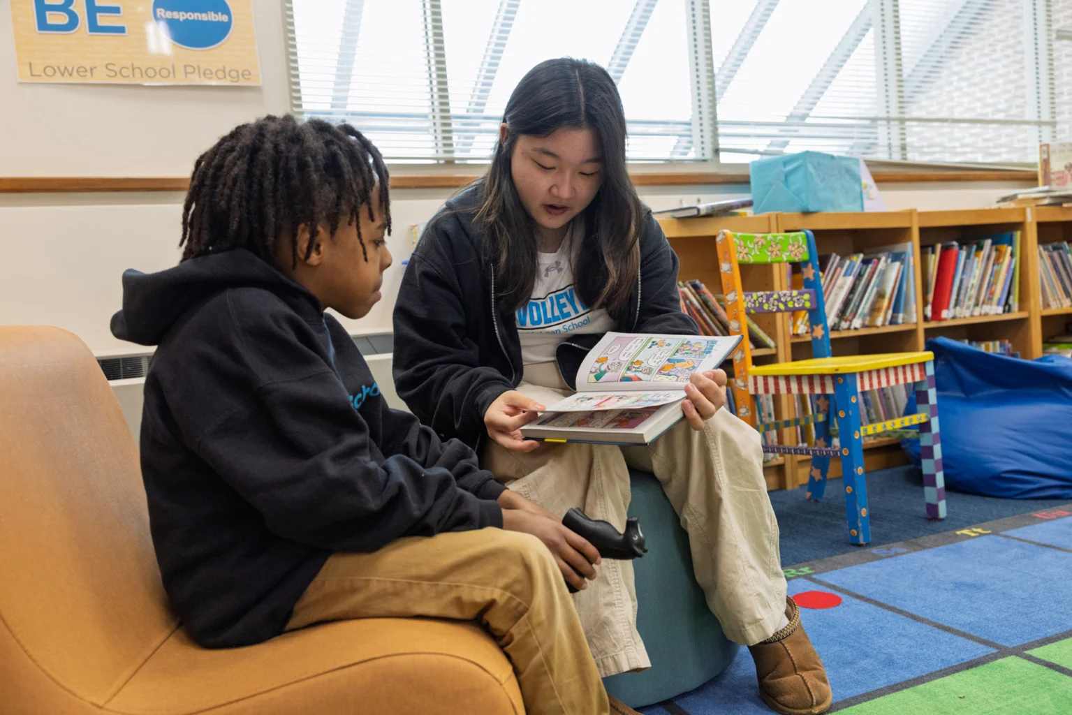 A teenage girl reads a comic book to a younger boy in a library, both seated on chairs with bookshelves and children's books visible in the background.