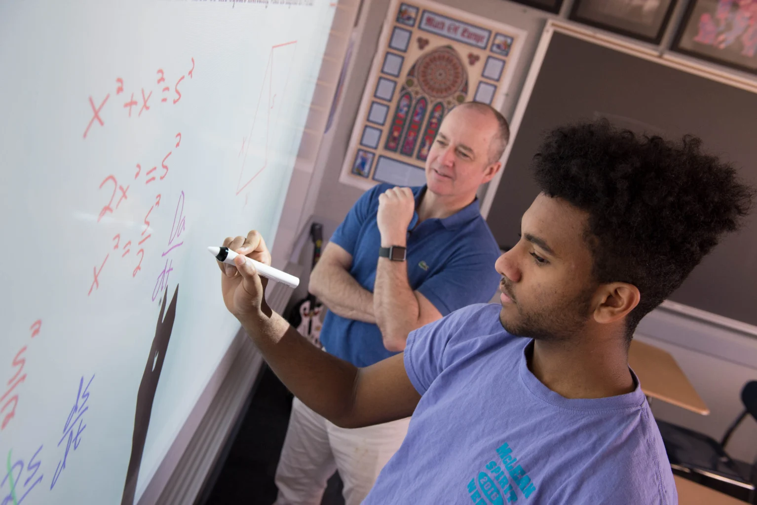 A student writes algebraic equations on a whiteboard while a teacher observes in a classroom setting.