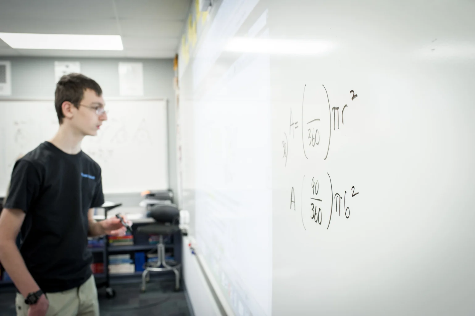 A student stands at a whiteboard with mathematical equations written on it in a classroom setting.