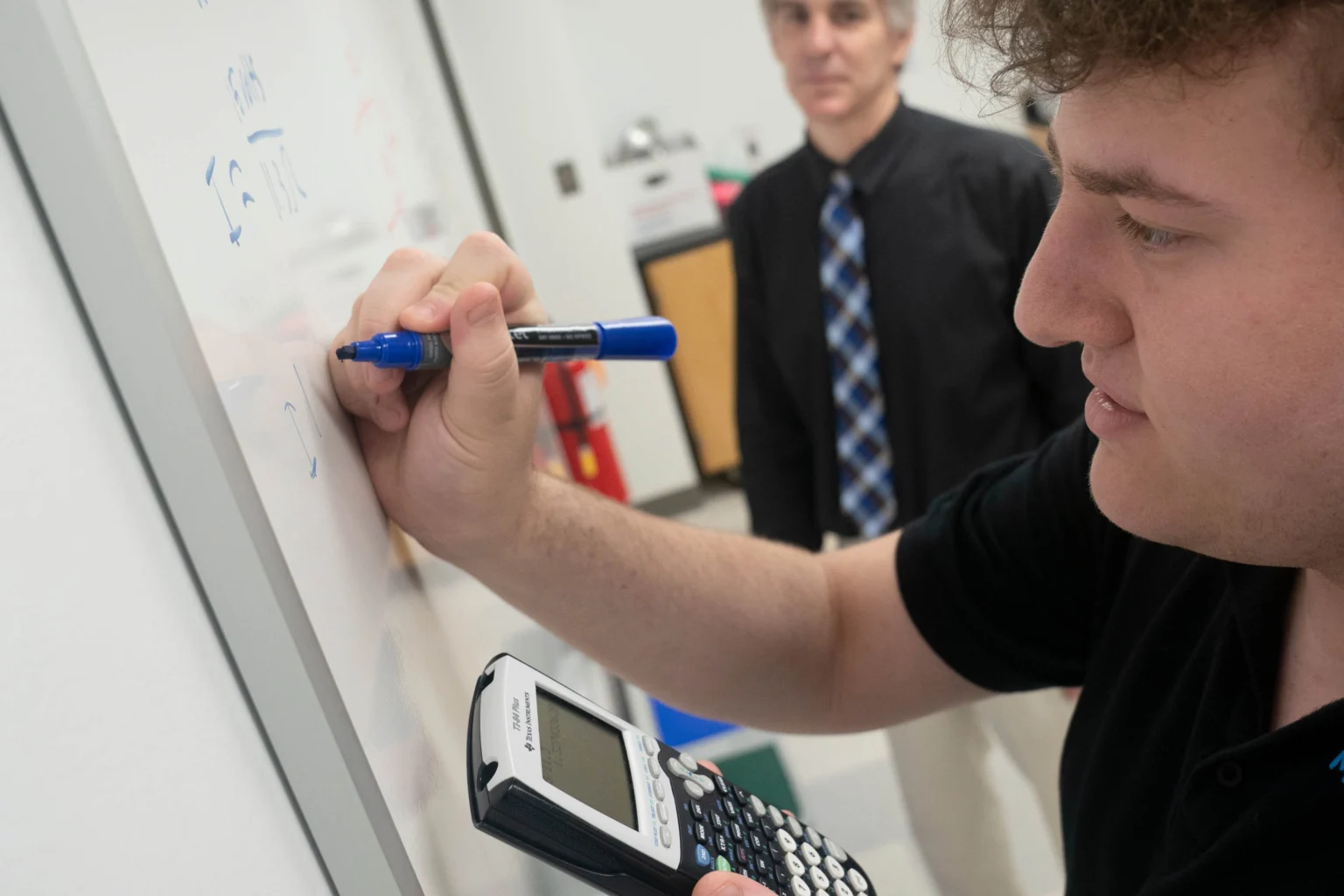 A person writes a math equation on a whiteboard with a marker while holding a calculator; another person stands in the background.